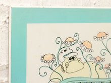 Load image into Gallery viewer, A close-up of a framed or mounted art print showing a whimsical cartoon illustration of a beige character wearing a white cap, surrounded by peach and white flowers with teal stems, against a white textured wall.
