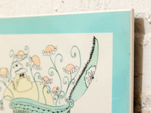 Load image into Gallery viewer, A close-up of a framed or mounted art print showing a whimsical illustration of a yellow creature in a teal bowl with peach flowers, against a white wall.
