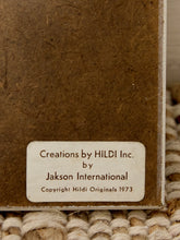 Load image into Gallery viewer, Close-up of a textured brown book or album cover with a cream label reading 'Creations by HILDI Inc. by Jakson International Copyright Hildi Originals 1973,' resting on a woven beige rug.
