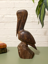 Load image into Gallery viewer, A dark brown carved wooden pelican sculpture perched on a chunky tree stump base, displayed on a sage green tabletop against a white brick wall with a green plant leaf in the background.
