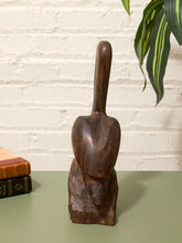 Load image into Gallery viewer, A dark brown carved wooden sculpture of a stylized face with a long handle, displayed on a green tabletop against a white brick wall background.
