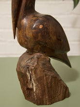 Load image into Gallery viewer, A dark brown carved wooden sculpture of a bird perched on a rough tree stump base, photographed indoors against a light wall and muted green surface.
