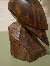 Load image into Gallery viewer, A dark brown carved wooden sculpture of a bird perched on a rough tree stump base, photographed indoors against a muted green surface and light neutral background.
