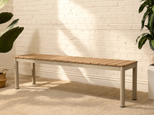 Load image into Gallery viewer, A modern outdoor bench with a light brown composite wood slat seat and a light gray metal frame, positioned against a white brick wall with natural sunlight casting shadows.
