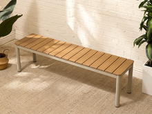 Load image into Gallery viewer, A modern outdoor bench with a light brown composite wood slatted seat and light gray metal legs, positioned on a beige carpeted floor against a white brick wall with potted plants.
