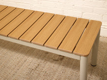 Load image into Gallery viewer, A modern outdoor bench with a light brown composite wood slatted seat and a white metal frame, shown on a woven rug against a white brick wall.
