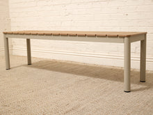 Load image into Gallery viewer, A modern outdoor bench with a light brown wooden slatted seat and a light gray frame, shown on a textured beige rug against a white brick wall.

