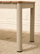 Load image into Gallery viewer, Close-up of a table leg in a warm off-white finish with a light natural wood top, standing on a beige woven rug against a white brick wall.
