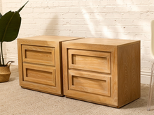 Load image into Gallery viewer, Two light oak wood nightstands with two drawers each, positioned side-by-side on a beige carpet against a white brick wall.

