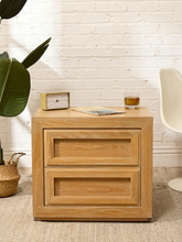 Load image into Gallery viewer, A light oak wood two-drawer nightstand with a mid-century modern design, styled in a room setting with a white brick wall, a potted plant, and a white chair.
