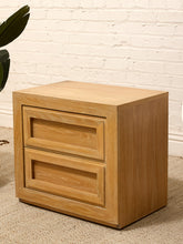 Load image into Gallery viewer, A light oak wood two-drawer nightstand or end table with a warm honey finish, set against a white brick wall and beige carpet.
