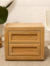 Load image into Gallery viewer, A light oak wood two-drawer nightstand with a warm honey finish, set against a white brick wall background with a green plant and a white chair partially visible.
