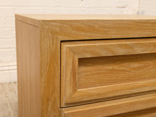 Load image into Gallery viewer, Close-up of a light oak wood chest of drawers showing the drawer front, side panel, and wood grain texture against a white brick wall background.

