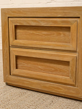 Load image into Gallery viewer, Close-up of a light oak wood two-drawer cabinet or nightstand with recessed panel doors, standing on a textured beige rug against a white brick wall.
