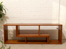 Load image into Gallery viewer, A mid-century modern wooden console table with a warm brown finish, featuring open shelving and a bench-style seat, set against a white brick wall.

