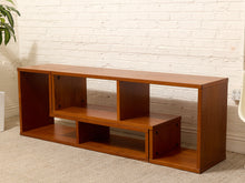 Load image into Gallery viewer, A wooden media console shelf with multiple open cubbies, shown in a warm medium-brown finish against a white brick wall and light carpet.
