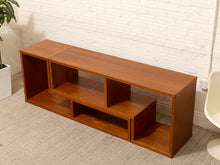 Load image into Gallery viewer, A mid-century modern wooden TV stand with a warm brown finish, featuring open cubbies and a lower pull-out shelf, set against a white brick wall and beige carpet.
