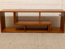 Load image into Gallery viewer, A mid-century modern wooden media console with a warm brown finish, featuring open shelving and a lower bench section, set against a white brick wall and textured beige carpet.
