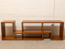 Load image into Gallery viewer, A mid-century modern wooden media console with a warm brown finish, featuring multiple open shelves and a geometric design, set against a white brick wall and light beige carpet.
