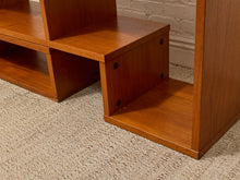 Load image into Gallery viewer, Close-up of a mid-century modern wooden shelving unit with a warm medium-brown finish, featuring open cubbies and open side shelves, set on a textured beige carpet against a white brick wall.
