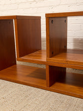 Load image into Gallery viewer, A close-up of a mid-century modern wooden shelving unit with a warm medium-brown finish, featuring open cubby compartments, set against a white brick wall and textured beige carpet.
