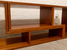 Load image into Gallery viewer, A mid-century modern wooden shelving unit with a warm reddish-brown finish, featuring open cubby shelves, set against a white brick wall and light-colored carpet.
