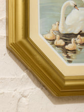 Load image into Gallery viewer, Close-up of a framed art print featuring a white swan and yellow ducklings in water, with a textured gold frame and cream mat against a white brick wall.
