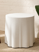 Load image into Gallery viewer, A white round tablecloth draped over a cylindrical side table, set against a white brick wall and beige carpet.
