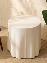 Load image into Gallery viewer, A round white fabric-covered stool or ottoman with a scalloped flared base, shown in a styled interior setting against a white brick wall on a woven rug.

