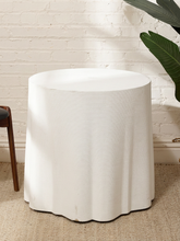 Load image into Gallery viewer, A white fabric-covered cylindrical side table or stool with a scalloped hem, shown in a styled room setting against a white brick wall with a green plant leaf in the background.
