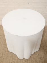 Load image into Gallery viewer, A white cylindrical side table or stool with a scalloped flared base, shown on a woven jute rug against a white brick wall.
