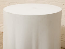 Load image into Gallery viewer, A white cylindrical table skirt or pedestal cover made of textured fabric, shown draped over a round table against a light brick wall background.
