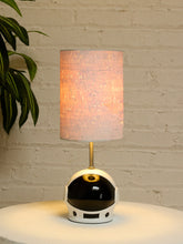Load image into Gallery viewer, A table lamp shaped like a white astronaut helmet with a black visor and gold accents, topped with a glowing warm beige fabric drum shade, set on a white surface against a white brick wall with a green plant leaf at the left edge.

