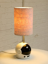 Load image into Gallery viewer, A table lamp shaped like an astronaut helmet with a glowing cylindrical shade, set on a white surface against a white brick wall.

