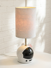 Load image into Gallery viewer, A table lamp with a cylindrical textured white shade and a base shaped like a white astronaut helmet with a black visor, shown lit on a white surface against a white brick wall.
