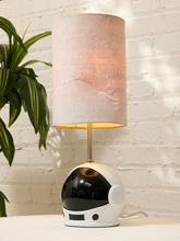 Load image into Gallery viewer, A table lamp with a white astronaut helmet base and a textured beige drum shade, set against a white brick wall with a green plant on the left.
