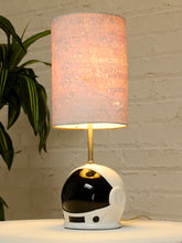 Load image into Gallery viewer, A table lamp with a cylindrical drum shade in a textured light pink fabric, mounted on a gold-finished stem. The base is shaped like a glossy white astronaut helmet with a black visor.
