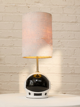 Load image into Gallery viewer, A table lamp shaped like an astronaut helmet with a glowing cylindrical shade, set against a white brick wall background.
