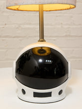 Load image into Gallery viewer, A table lamp shaped like a white astronaut helmet with a glossy black visor, a gold-toned stem, and a light beige lampshade, set against a white brick wall background.
