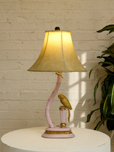 Load image into Gallery viewer, A pink and gold table lamp featuring a curved bamboo-style stem with a gold bird perched on it, set against a white brick wall and a green plant.
