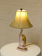 Load image into Gallery viewer, A decorative table lamp with a glowing warm yellow shade, a curved pink stem base, and a gold parrot figurine perched on the side, set on a white round table against a white brick wall.
