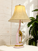 Load image into Gallery viewer, A pink table lamp with a curved bamboo-style stem base, gold-toned decorative accents, and a beige fabric bell shade, shown on a white surface against a white brick wall with a green plant nearby.
