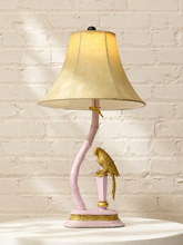 Load image into Gallery viewer, A decorative table lamp with a warm beige fabric shade, a curved pink base, and a gold bird figurine perched on the side, set against a white brick wall.
