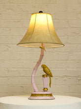 Load image into Gallery viewer, A decorative table lamp with a glowing cream-colored bell-shaped shade, a curved pale pink base, and two gold bird figurines perched on the structure, set against a white brick wall.

