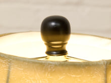 Load image into Gallery viewer, Close-up of a table lamp shade and finial, showing a warm cream fabric shade with a glossy black round finial under glowing light.
