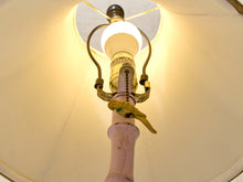 Load image into Gallery viewer, A close-up, low-angle view of a vintage-style table lamp with a light pink ceramic base, gold-toned metal arms, and a glowing white glass bulb inside a cream-colored shade.
