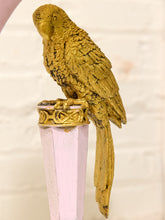 Load image into Gallery viewer, A gold-painted parrot figurine perched on top of a pale pink decorative finial, photographed against a softly blurred light background.

