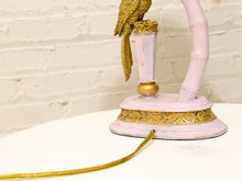 Load image into Gallery viewer, A close-up of a decorative table lamp with a pale pink bamboo-style curved arm, a gold bird figurine perched on the top, and ornate gold trim on a round pink base, set against a white brick wall.
