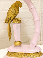 Load image into Gallery viewer, A decorative sculpture featuring a textured gold parrot perched on a pale pink cylindrical pedestal, with a curved pale pink bamboo-like arm and a round pink-and-gold base against a white brick wall background.
