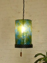 Load image into Gallery viewer, A hanging pendant light with a cylindrical glass shade featuring a mottled green and blue pattern, illuminated from within. The shade is finished with a black metal band and a hanging pull chain with a tassel, set against a white brick wall background.
